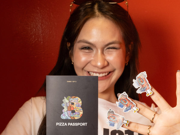 Smiling woman in a Ballistic Pizza tee holding Pizza Passport Card 1, with holographic PIM stickers on her fingers